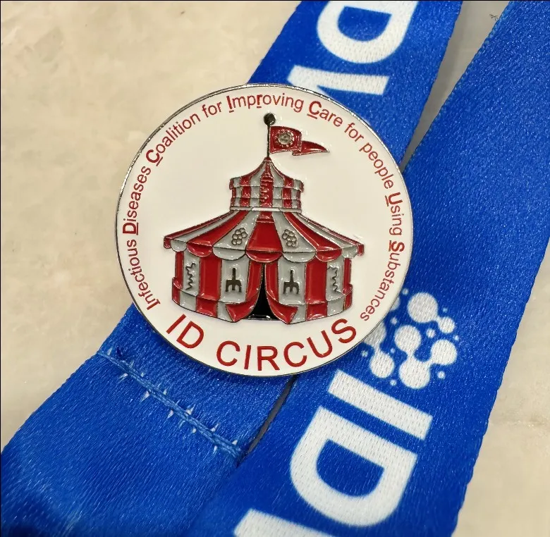 An enamel pin on a conference lanyard: a red and white big top tent, ringed by the words Infectious Diseases Coalition for Improving Care for people Using Substances, with ID CIRCUS across the bottom.